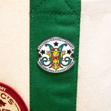 Load image into Gallery viewer, A close-up of a circular enamel pin featuring a stylized 'MM' monogram with the text 'READING TERMINAL MARKET' and 'PHILADELPHIA • PENNSYLVANIA' on a green fabric strip against a cream canvas background.
