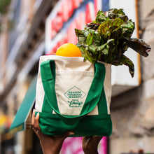 Load image into Gallery viewer, A person holds a cream canvas tote bag with green handles and a green printed logo reading 'Lea’ning Farmers Market Philadelphia' while carrying leafy greens and a yellow fruit in a blurred outdoor street setting.
