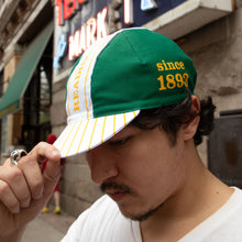 Load image into Gallery viewer, A man wearing a green and white baseball cap with yellow stripes and yellow text reading 'since 1893' on a city street.
