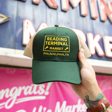 Load image into Gallery viewer, A hand holds a green and beige trucker hat with a yellow embroidered patch reading 'READING TERMINAL MARKET PHILADELPHIA, PA' in front of a colorful market sign.
